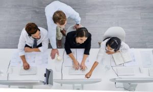 Team of IT help desk professionals working at a row of computers, collaborating and assisting each other while wearing headsets in a modern office.