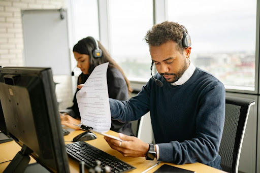IT support staff wearing headsets working at office desks, with one agent reviewing printed documents while using a computer.