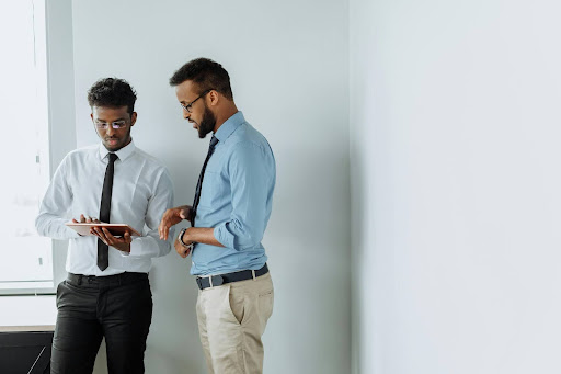 Two professionals standing in an office reviewing information on a tablet, discussing work in a bright, minimal workspace.