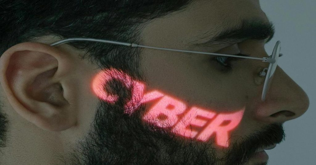 Close-up side profile of a man wearing glasses with the word ‘CYBER’ projected in red light across his beard and cheek.